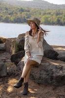 A portrait of a beautiful woman wearing a hat sitting on a rock against a beautiful mountain and water source. photo