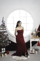 Portrait of a pretty young girl wearing a red gown, smiling at the camera, standing in decorated Christmas living room indoors photo