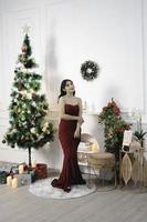 Portrait of a pretty young girl wearing a red gown, smiling at the camera, standing in decorated Christmas living room indoors photo