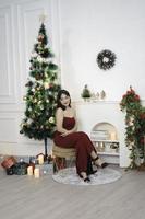 Portrait of pretty young girl cozy sit down, smiling wear red gown in decorated Christmas living room indoors photo