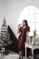 retrato de una linda joven con un vestido rojo y bebiendo vino, sonriendo a la cámara, de pie en un salón de Navidad decorado en el interior foto