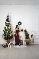 Portrait of a pretty young girl wearing a red gown, smiling at the camera, standing in decorated Christmas living room indoors photo