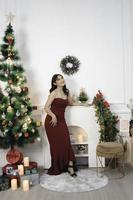 Portrait of a pretty young girl wearing a red gown, smiling at the camera, standing in decorated Christmas living room indoors photo