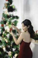 Portrait of pretty young girl decorating Christmas tree, smiling wearing red gown in decorated Christmas living room indoors photo