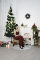 Portrait of pretty young girl cozy sit down, smiling wear red gown in decorated Christmas living room indoors photo