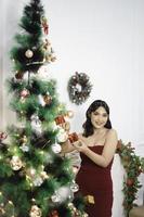 Portrait of pretty young girl decorating Christmas tree, smiling wearing red gown in decorated Christmas living room indoors photo