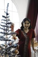 Portrait of pretty young girl decorating Christmas tree, smiling wearing red gown in decorated Christmas living room indoors photo