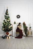 Portrait of pretty young girl cozy sit down, smiling wear red gown in decorated Christmas living room indoors photo