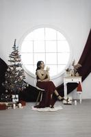 Portrait of pretty young girl cozy sit down and drinking wine, wear red gown in decorated Christmas living room indoors photo