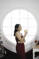 Portrait of a pretty young girl wearing a red gown and drinking wine, smiling at the camera, standing in decorated Christmas living room indoors photo