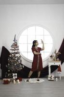 retrato de una linda joven con un vestido rojo, sonriendo a la cámara, de pie en un salón de Navidad decorado en el interior foto