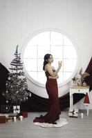 retrato de una linda joven con un vestido rojo y bebiendo vino, sonriendo a la cámara, de pie en un salón de Navidad decorado en el interior foto