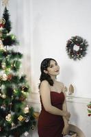 Portrait of a pretty young girl wearing a red gown, smiling at the camera, standing in decorated Christmas living room indoors photo