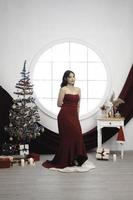 Portrait of a pretty young girl wearing a red gown, smiling at the camera, standing in decorated Christmas living room indoors photo