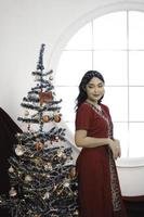 retrato de una linda joven decorando un árbol de navidad, sonriendo usando un vestido rojo en una sala decorada de navidad en el interior foto