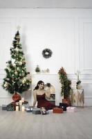 Portrait of pretty young girl cozy sit down holding Christmas present, smiling wear red gown in decorated Christmas living room indoors photo