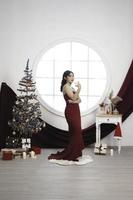 retrato de una linda joven con un vestido rojo y bebiendo vino, sonriendo a la cámara, de pie en un salón de Navidad decorado en el interior foto