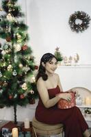 Portrait of pretty young girl cozy sit down holding Christmas present, smiling wear red gown in decorated Christmas living room indoors photo