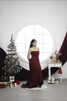 Portrait of a pretty young girl wearing a red gown, smiling at the camera, standing in decorated Christmas living room indoors photo