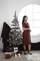 Portrait of a pretty young girl wearing a red gown, smiling at the camera, standing in decorated Christmas living room indoors photo