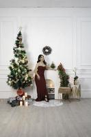 retrato de una linda joven con un vestido rojo, sonriendo a la cámara, de pie en un salón de Navidad decorado en el interior foto