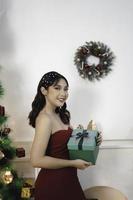 retrato de una joven bonita y acogedora sentada sosteniendo un regalo de navidad, vistiendo un vestido rojo sonriente en una sala de estar navideña decorada en el interior foto
