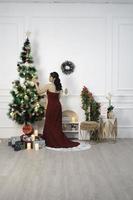 Portrait of pretty young girl decorating Christmas tree, smiling wearing red gown in decorated Christmas living room indoors photo