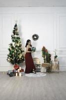 Portrait of pretty young girl cozy sit down holding Christmas present, smiling wear red gown in decorated Christmas living room indoors photo