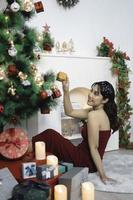 Portrait of pretty young girl cozy sit down holding Christmas present, smiling wear red gown in decorated Christmas living room indoors photo