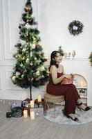 Portrait of pretty young girl cozy sit down holding Christmas present, smiling wear red gown in decorated Christmas living room indoors photo