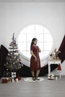 Portrait of a pretty young girl wearing a red gown, smiling at the camera, standing in decorated Christmas living room indoors photo