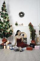 Portrait of pretty young girl cozy sit down holding Christmas present, smiling wear red gown in decorated Christmas living room indoors photo