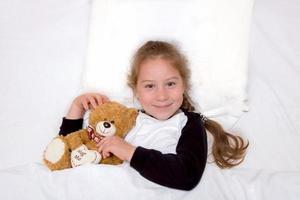 girl lies in bed with a bear and smiles photo