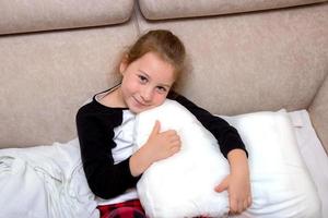 girl after an illness in pajamas sits on a bed hugs a pillow and smiles photo