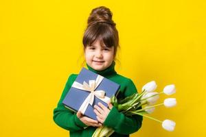 a happy little brunette girl is holding a gift box and a bouquet of tulips on a yellow background in the studio. The concept of Mother's Day, March 8, Valentine's Day. High quality photo