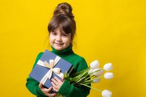 a happy little brunette girl is holding a gift box and a bouquet of tulips on a yellow background in the studio. The concept of Mother's Day, March 8, Valentine's Day. High quality photo