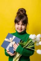 a happy little brunette girl is holding a gift box and a bouquet of tulips on a yellow background in the studio. The concept of Mother's Day, March 8, Valentine's Day. High quality photo