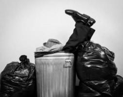 Black and White Portrait of Businessman in Suit Sitting in Metal Trash Can Surrounded by Trash Bags.  Concept of Man Beaten by Capitalism and Life. photo
