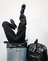 hombre de negocios con traje atrapado boca abajo en un bote de basura de metal junto a la pila de bolsas de basura. concepto de más de un barril. desechados por el capitalismo y la codicia.