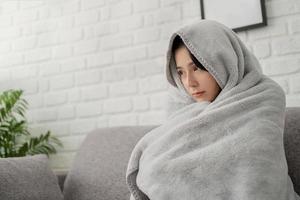 Sick young asian woman sit on sofa at home wrapped covered in warm duvet or blanket, She feeling unhealthy ill. photo