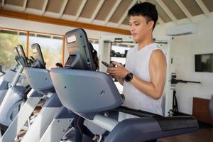 joven asiático usando un teléfono inteligente en una caminadora en el gimnasio. foto