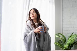 Beautiful Asian woman wrapped in blanket leaning against window and looking outside. She is drinking hot tea. photo