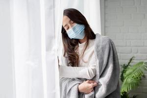Sick asian woman in medical face mask standing by the window and yearning to go outside, being on quarantine, ill with covid-19. Social distancing and quarantine concept