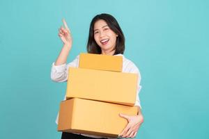 mujer asiática feliz sosteniendo una caja de paquetes y señalando con el dedo el espacio de copia vacío aislado en un fondo verde claro, foto