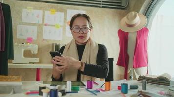 asiatisk medelålders kvinna mode designer Arbetar i studio förbi talande på mobil telefon handla om idéer och teckning skisser för klänning design samling order. professionell boutique skräddare sme entreprenör. video