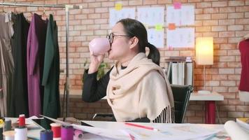 asiatische Modedesignerin mittleren Alters arbeitet in einem Kreativstudio, skizziert, trinkt Kaffee, denkt und zerkleinert Ideen in Kleiderdesign-Kollektionen und ist eine professionelle Boutique-Schneiderunternehmerin. video