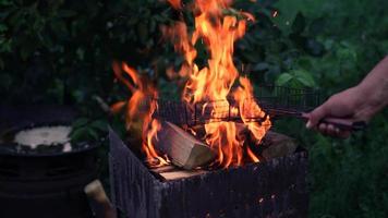 la mano del hombre calienta una parrilla de barbacoa en un fuego para carne a la parrilla. el fuego arde en el brasero de la parrilla y saltan chispas. picnic nocturno en la naturaleza. carbón ardiendo con llamas y llamas. barbacoa de leña video