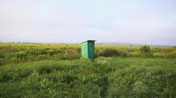 Alte Holztoilette mit einem geschnitzten Fenster mit der Form eines Herzens, das an der Tür ausgeschnitten ist, auf freiem Feld. Vintage-WC. Eine rustikale grüne Toilette im Freien in einer Feldlandschaft aus Blumen und Gras bei Sonnenaufgang video