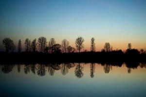 silhouette and reflection of trees by the lake at sunset photo