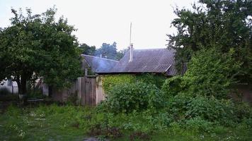wit rook is ontladen van een schoorsteen Aan de betegeld dak van een buitenwijk huis. damp gaan omhoog van de werf achter schutting. oud dorp huizen staan in een tuin tussen bomen en groen. video